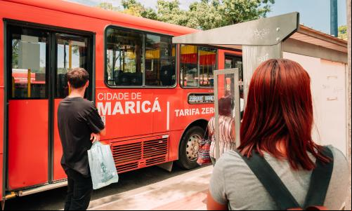 pessoas em ponto de ônibus e ônibus parado com escrito tarifa zero na lateral